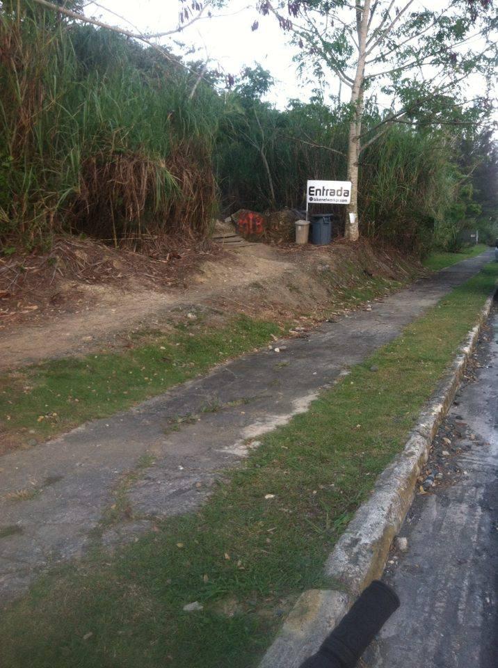 A dirt pathway merges with a concrete sidewalk beside a grassy area. On the left, there is a sign labeled "Entrada," indicating an entrance, along with a couple of trash bins. The pathway is surrounded by tall grasses and trees in the background. Monte Realengo mountain bike trail.