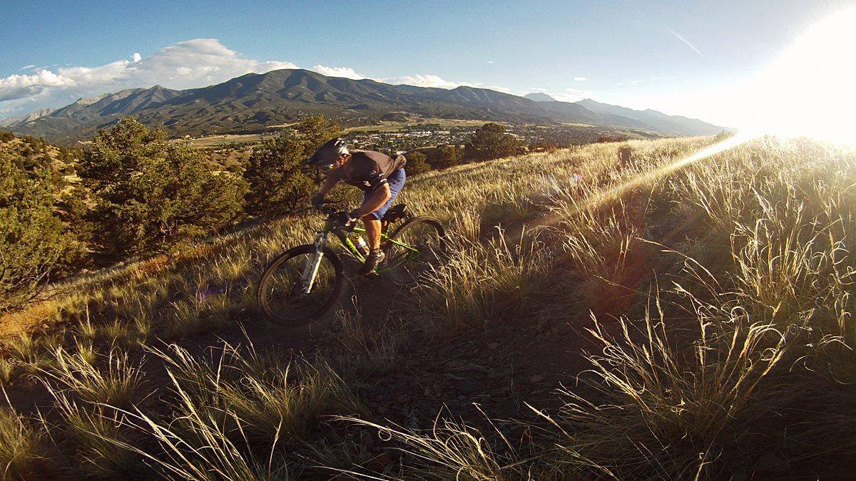 Airborne Goblin: A mountain biker riding on a grassy trail with a scenic view of mountains and a valley in the background, illuminated by sunlight.