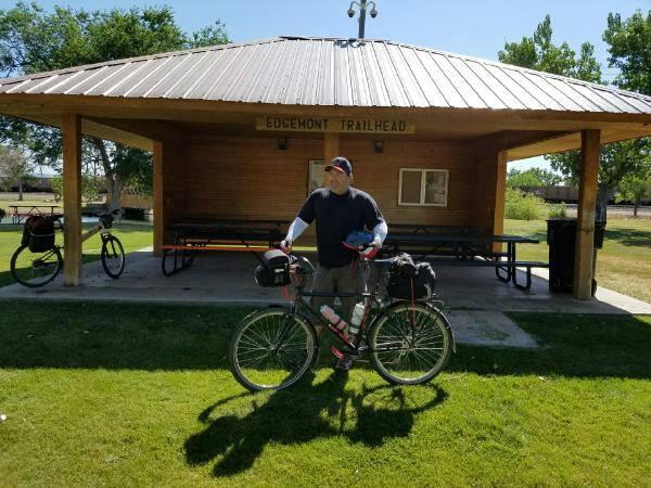 A person standing next to a bicycle in front of a park shelter marked "Edgemont Trailhead." The individual is casually dressed and holding a helmet, with another bicycle visible in the background on the grassy area. The scene is set in a sunny outdoor environment with trees lining the background. The George S. Mickelson Trail mountain bike trail.