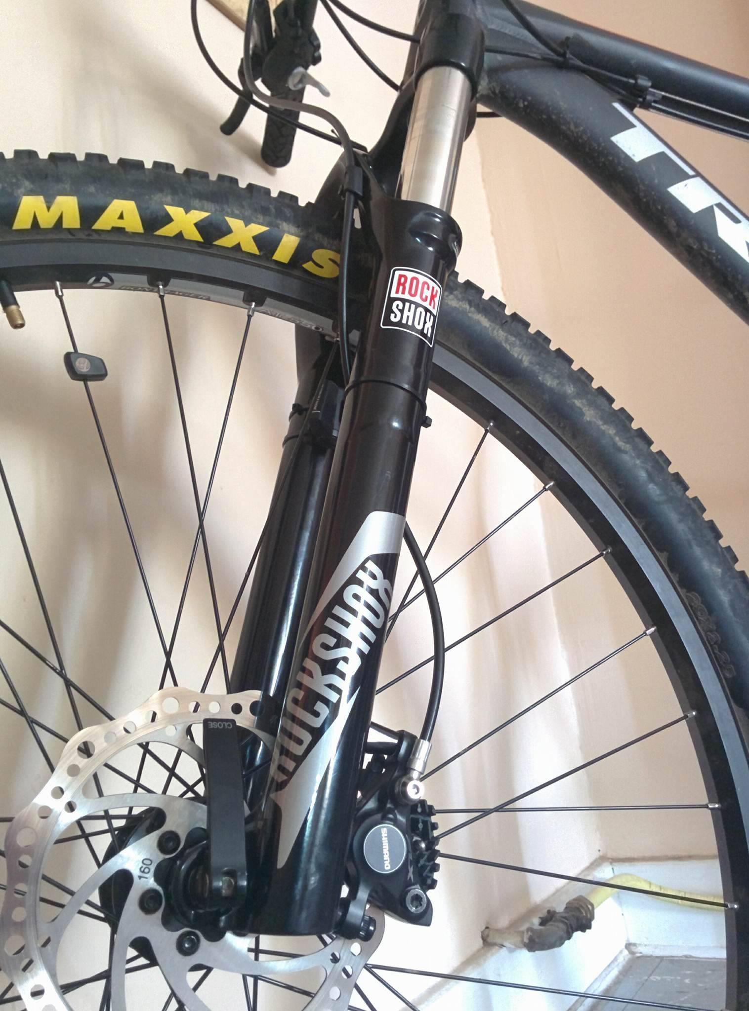 Trek Marlin 5 29er: Image of a mountain bike front fork featuring a black RockShox suspension system, with a Maxxis tire visible in the foreground. The wheel includes a silver disc brake and the bike is positioned against a wall.