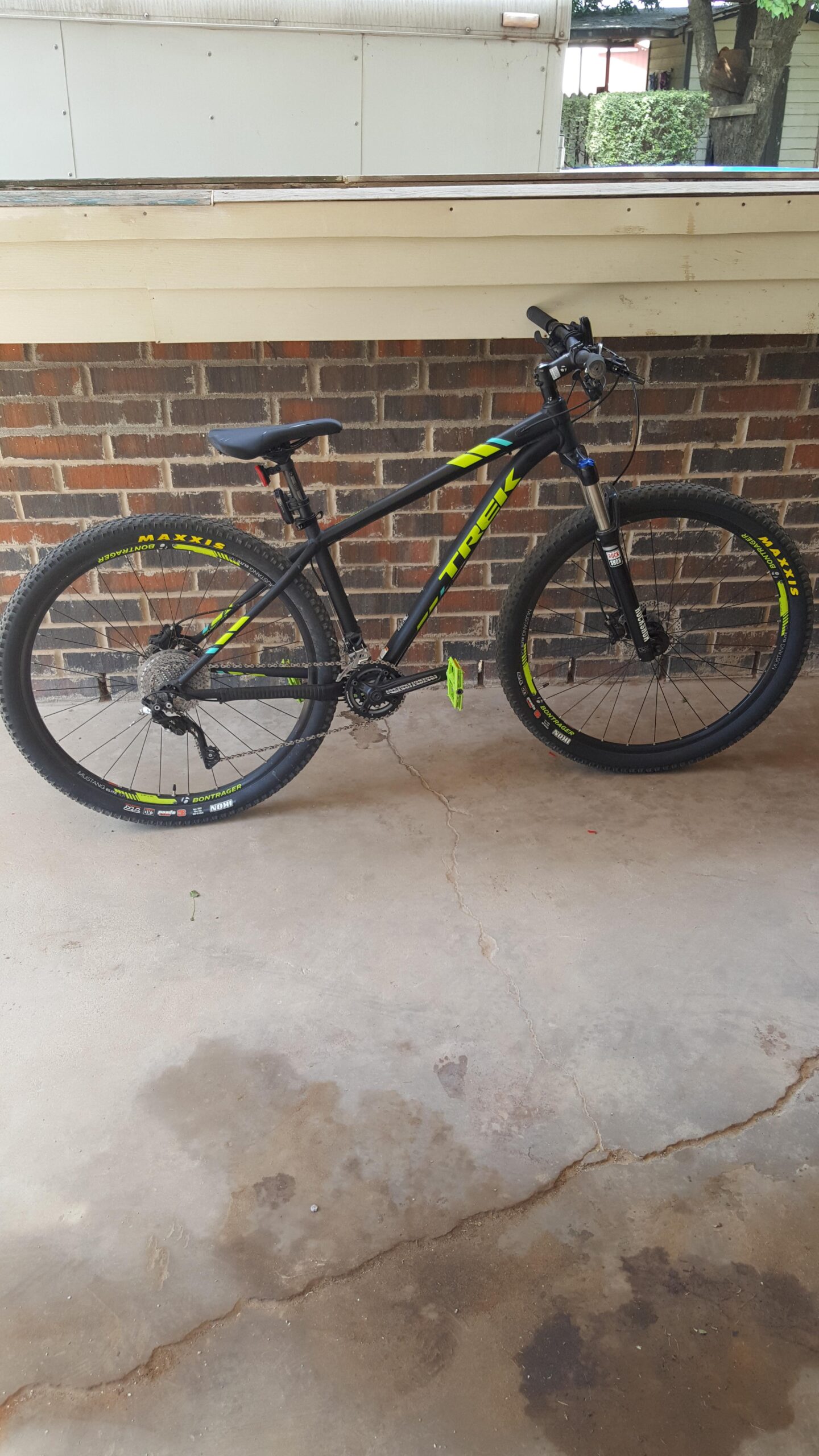 Trek X-Caliber 9: A black mountain bike with bright green accents parked on a concrete surface. The bike features thick tires and a sturdy frame, with visible gears and pedals. A brick wall is in the background, and some patches on the concrete floor are visible.