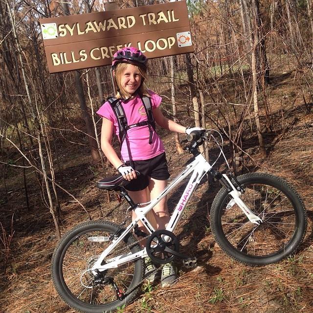 A young girl wearing a helmet and a pink shirt stands next to her bicycle at the Sylaward Trail and Bills Creek Loop sign, surrounded by trees and a natural setting. Sylaward mountain bike trail.