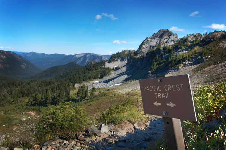 A scenic view of a mountainous landscape with a signpost labeled "Pacific Crest Trail," indicating directions to either side. The area is surrounded by lush green trees and rocky terrain under a clear blue sky. Mountains are visible in the distance, creating a tranquil and inviting atmosphere for hikers.