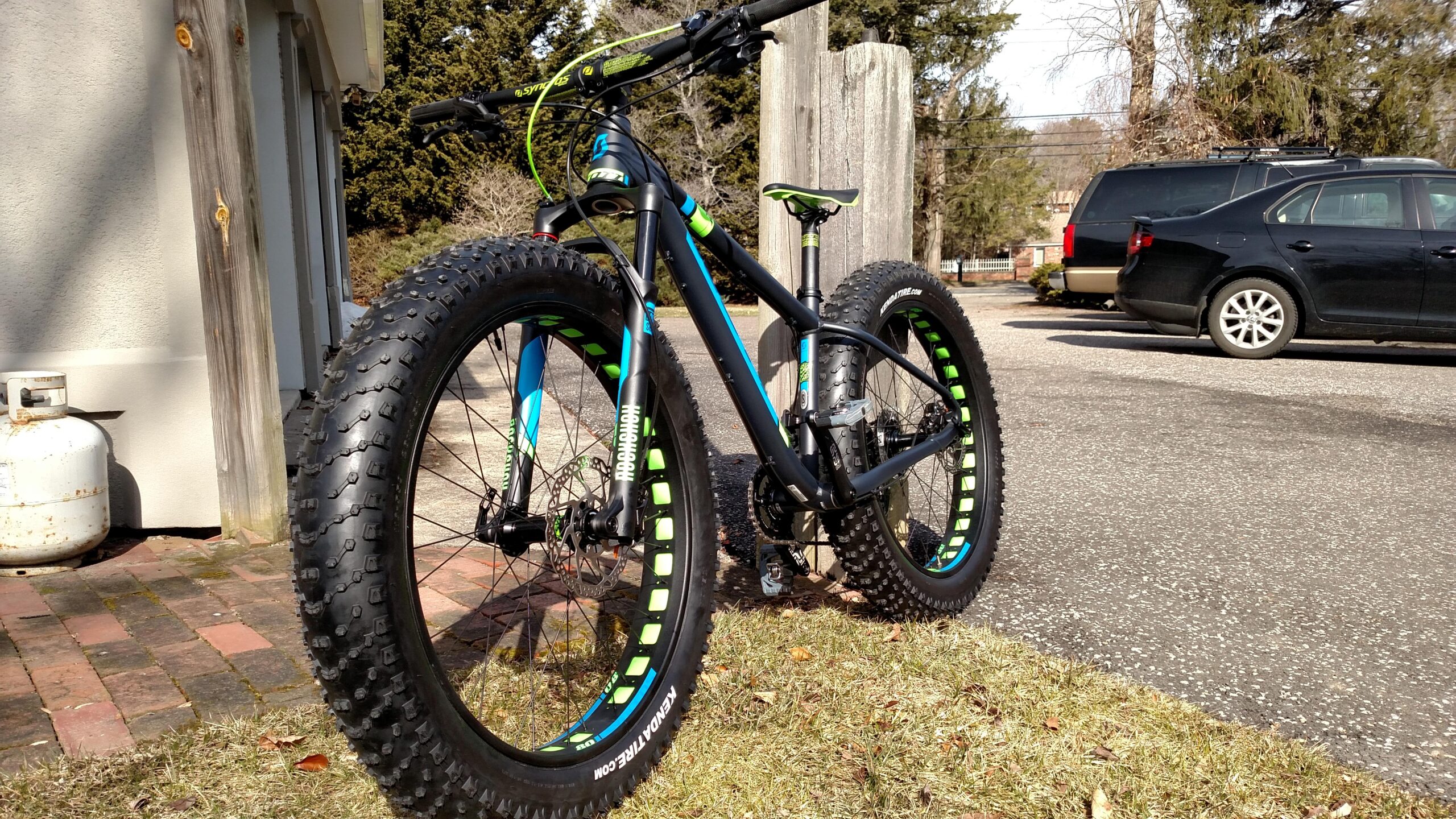 Scott Big Ed: A black fat bike with blue and green accents, parked on a driveway. The bike features large, knobby tires, a visible gear mechanism, and is positioned next to a wooden post. In the background, there are trees and parked vehicles.