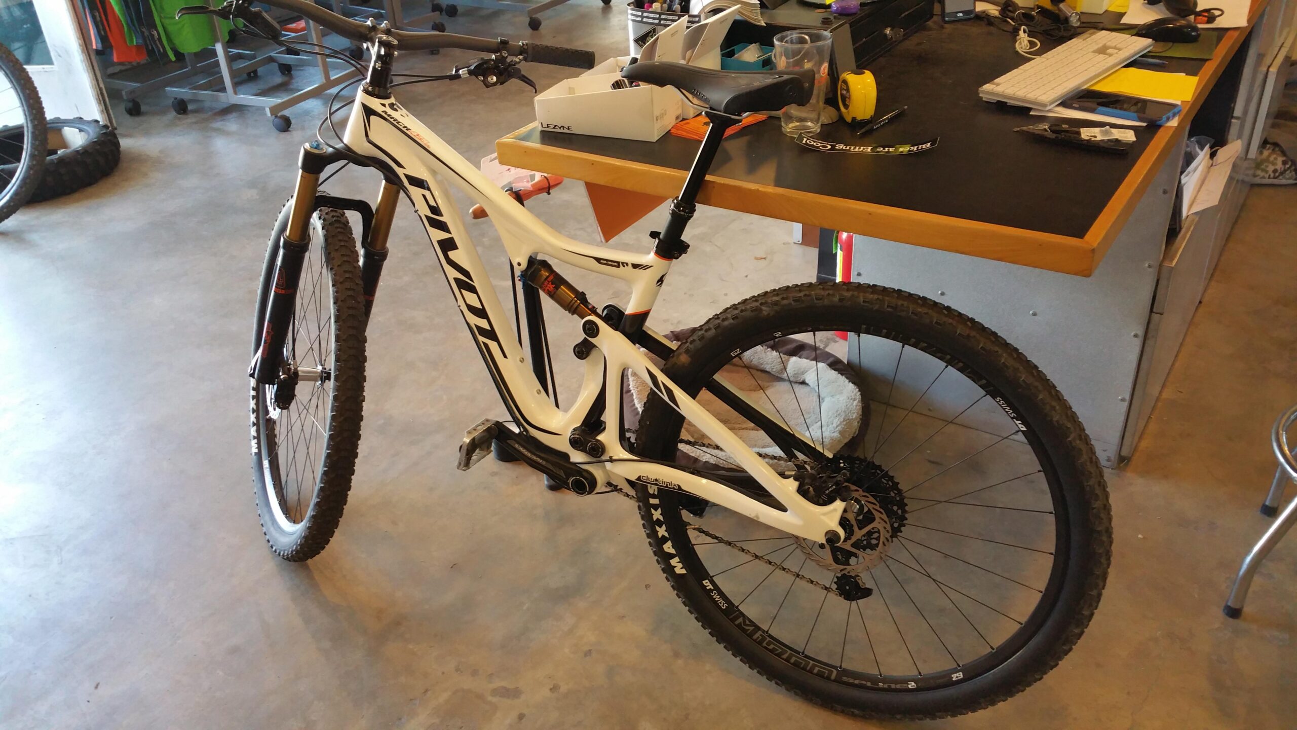 Pivot Trail 429: A mountain bike with a white frame and black wheels is positioned in a workshop. The bike features suspension forks and a dropper seat post. In the background, there are additional bicycles and various tools on a workbench, including a keyboard, measuring tape, and small containers. The floor is concrete, and there is a cozy mat near the bike.