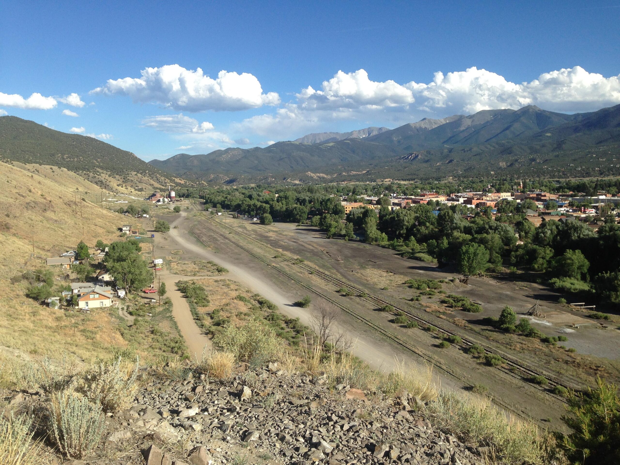 A scenic view of a valley with rolling hills and mountains in the background. In the foreground, a dirt road runs through a small area with scattered homes and trees. A town is visible in the distance, with colorful buildings among green foliage, under a blue sky dotted with fluffy white clouds. Arkansas Hills mountain bike trail.
