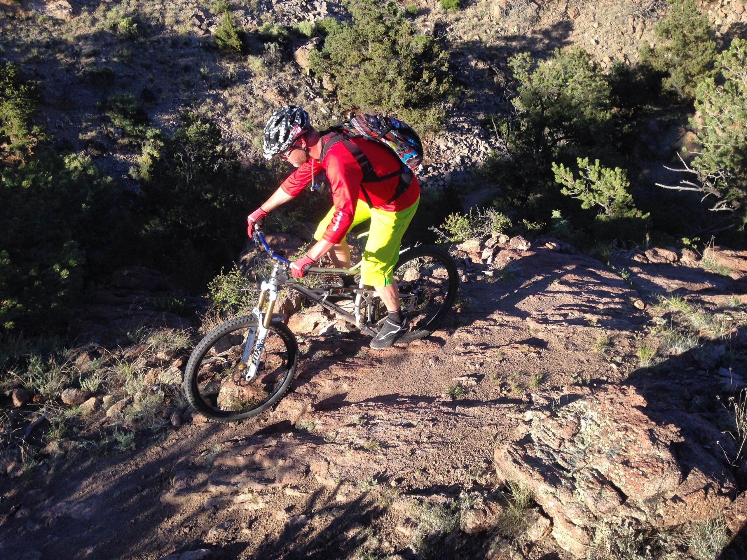 A mountain biker in a bright red shirt and yellow shorts navigates a rocky trail surrounded by greenery, showcasing an adventurous outdoor setting. Arkansas Hills mountain bike trail.