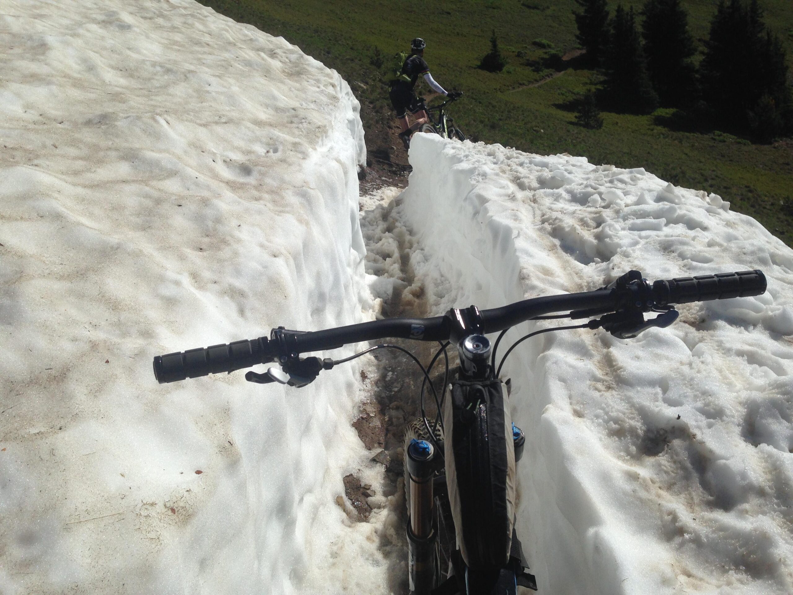 A mountain biker standing beside a snowy path with large snow banks on either side. The bike's handlebars are in the foreground, and another biker can be seen in the background navigating the snowy terrain. Lush green hills and trees are visible in the distance, indicating a mountainous landscape in late spring or early summer. Monarch Crest Trail mountain bike trail.