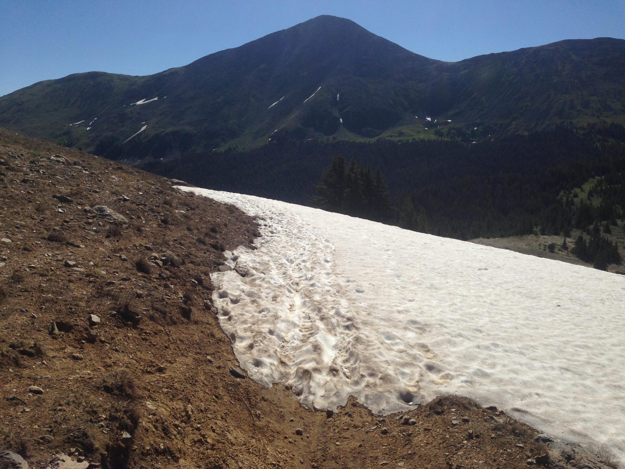 A mountainous landscape featuring a snow-covered slope on the right, transitioning to rocky and earthy terrain on the left. In the background, a prominent mountain peak rises against a clear blue sky. Sparse greenery is visible on the mountainside and in the valley below. Monarch Crest Trail mountain bike trail.