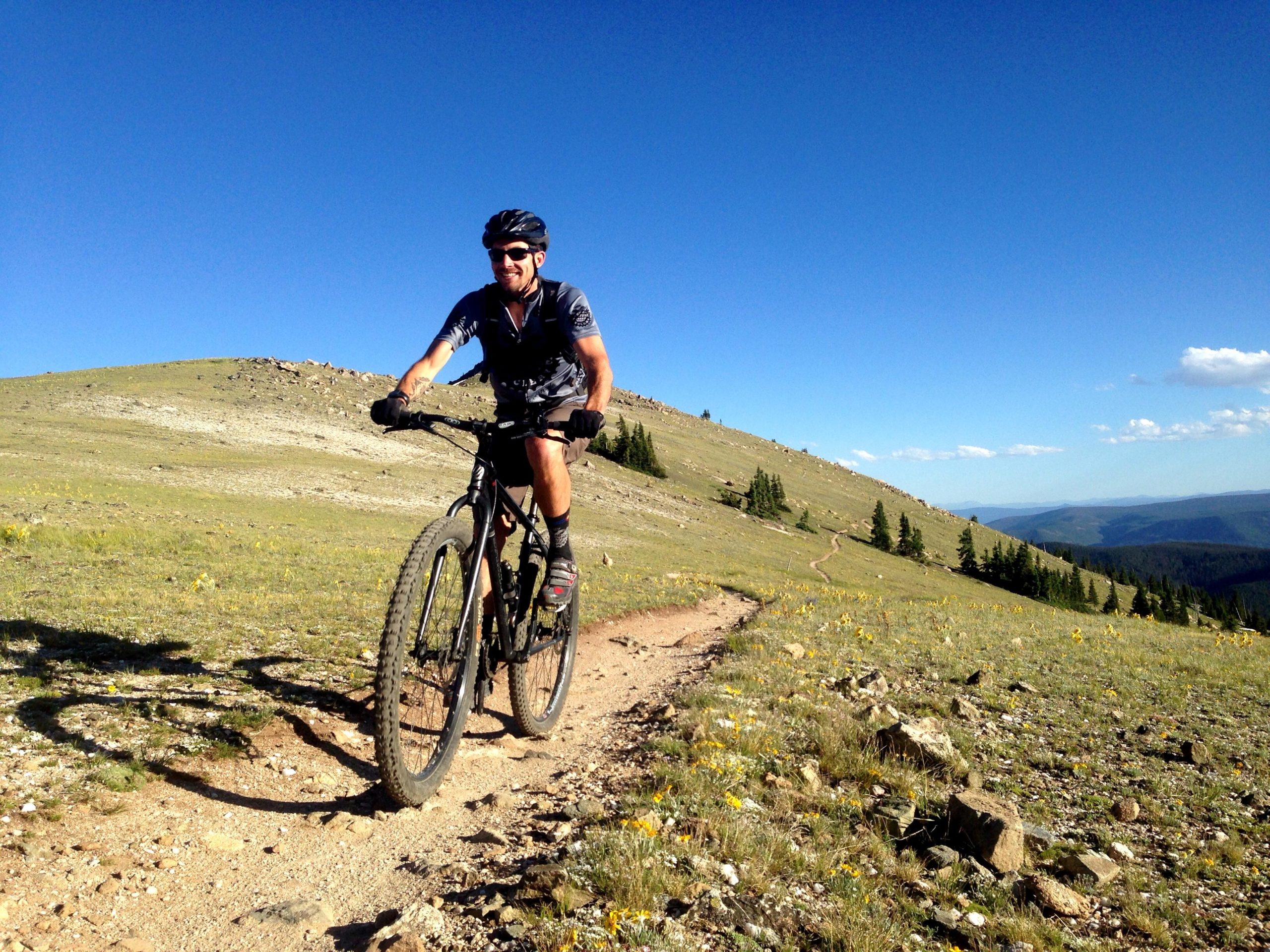 A mountain biker riding along a dirt trail on a green hillside under a clear blue sky. The cyclist is wearing a helmet and sunglasses, smiling as they navigate the terrain surrounded by trees and rocky formations in the background. Monarch Crest Trail mountain bike trail.