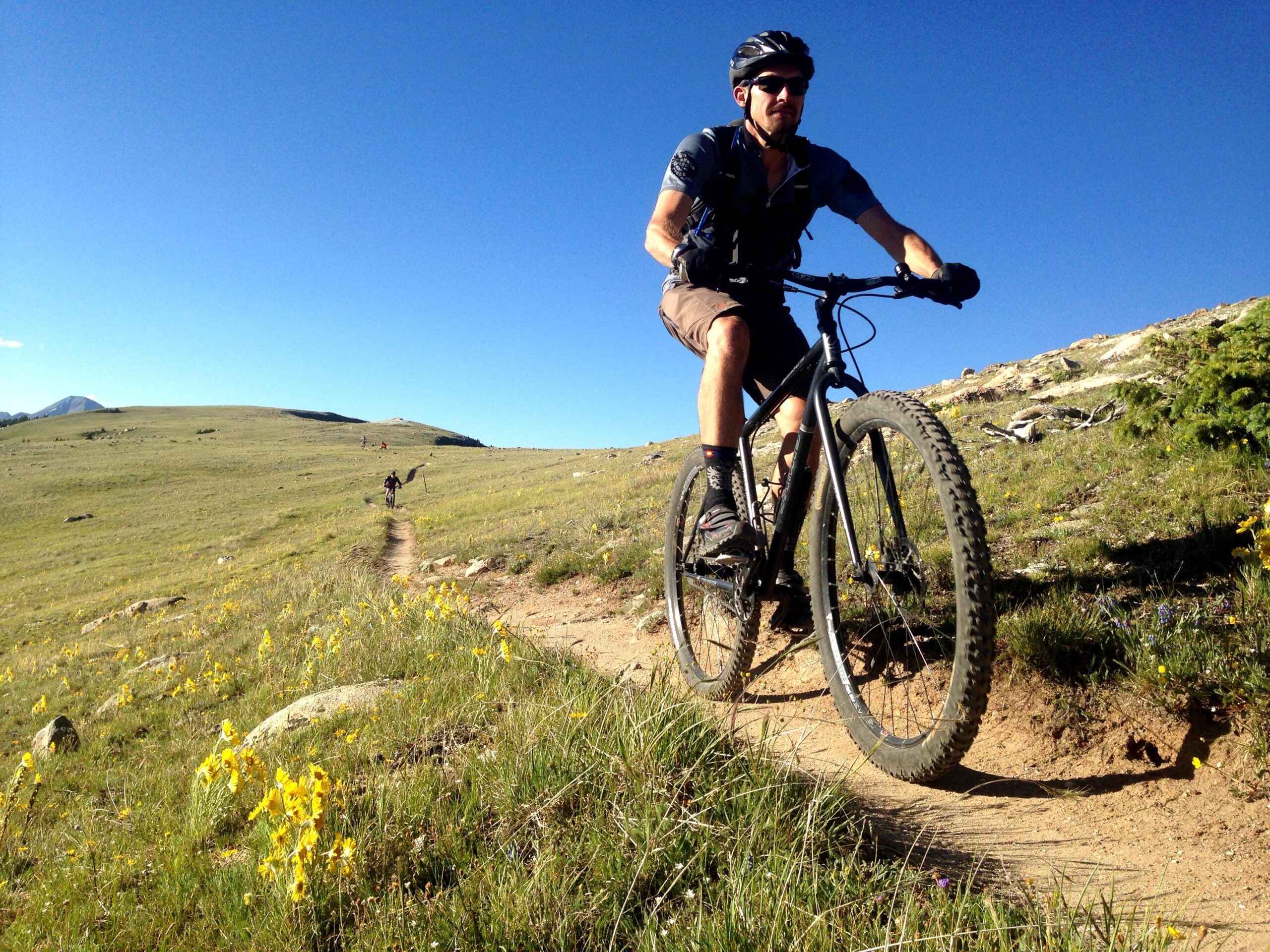 A person riding a mountain bike on a scenic trail surrounded by green grass and yellow wildflowers, with a clear blue sky overhead. In the background, other cyclists are visible on the trail, indicating a vibrant outdoor setting. Monarch Crest Trail mountain bike trail.