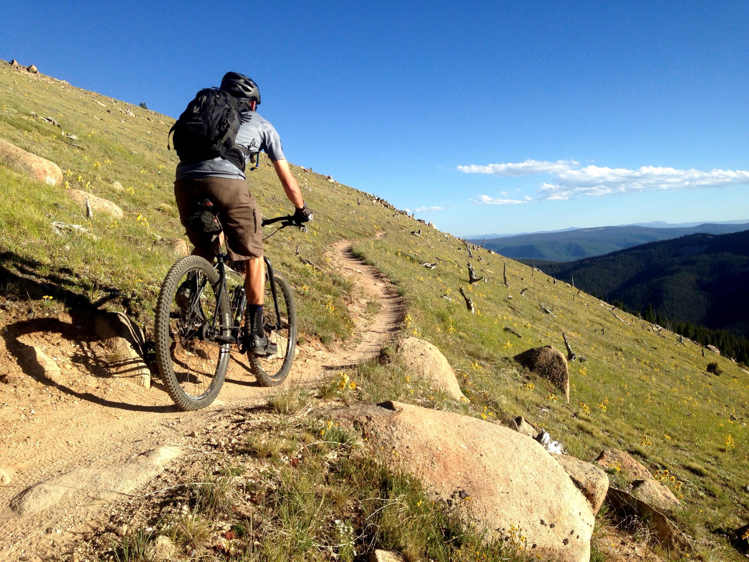 A person riding a mountain bike along a dirt trail on a hillside, surrounded by greenery and scattered rocks. The landscape features a clear blue sky and distant mountain ranges in the background. Monarch Crest Trail mountain bike trail.