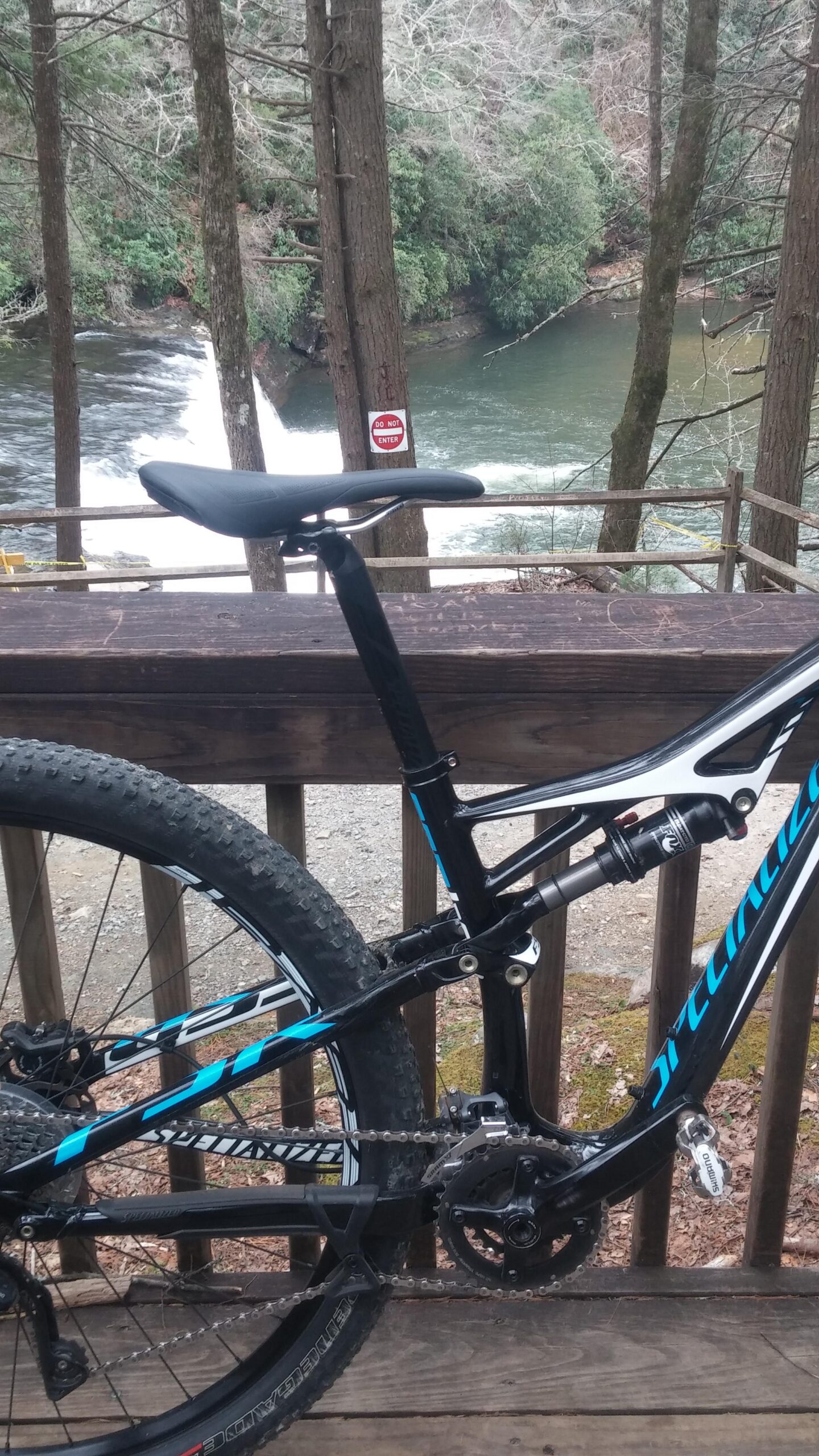 A stylish mountain bike leans against a wooden railing, with a view of a river and waterfall in the background. Signs warning not to enter are visible on a nearby tree, surrounded by lush greenery and a peaceful natural setting. DuPont State Forest mountain bike trail.