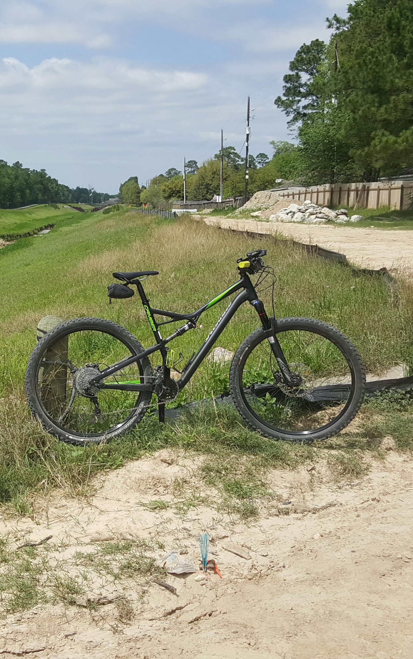 Specialized Camber Comp 29: A black and green mountain bike is parked on a gravel path beside a grassy area and a small waterway. In the background, there are trees and telephone poles, with a partially visible fence and a dirt road. The scene is bright and sunny with a few clouds in the sky.
