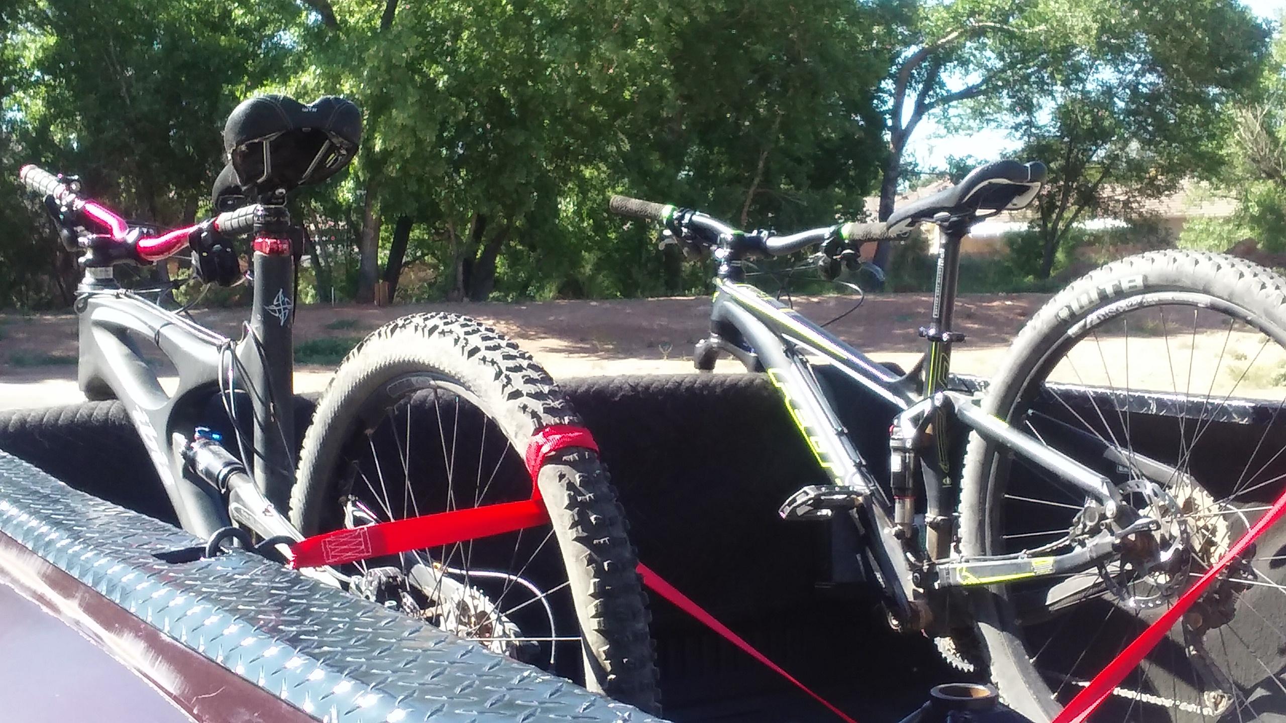 Felt Edict Nine 50: Two mountain bikes secured in the back of a pickup truck. One bike features a black frame with red accents, while the other is predominantly black with green accents. Both bikes have thick, knobby tires and are fastened with red straps against a textured black surface in the truck bed, surrounded by greenery in the background.