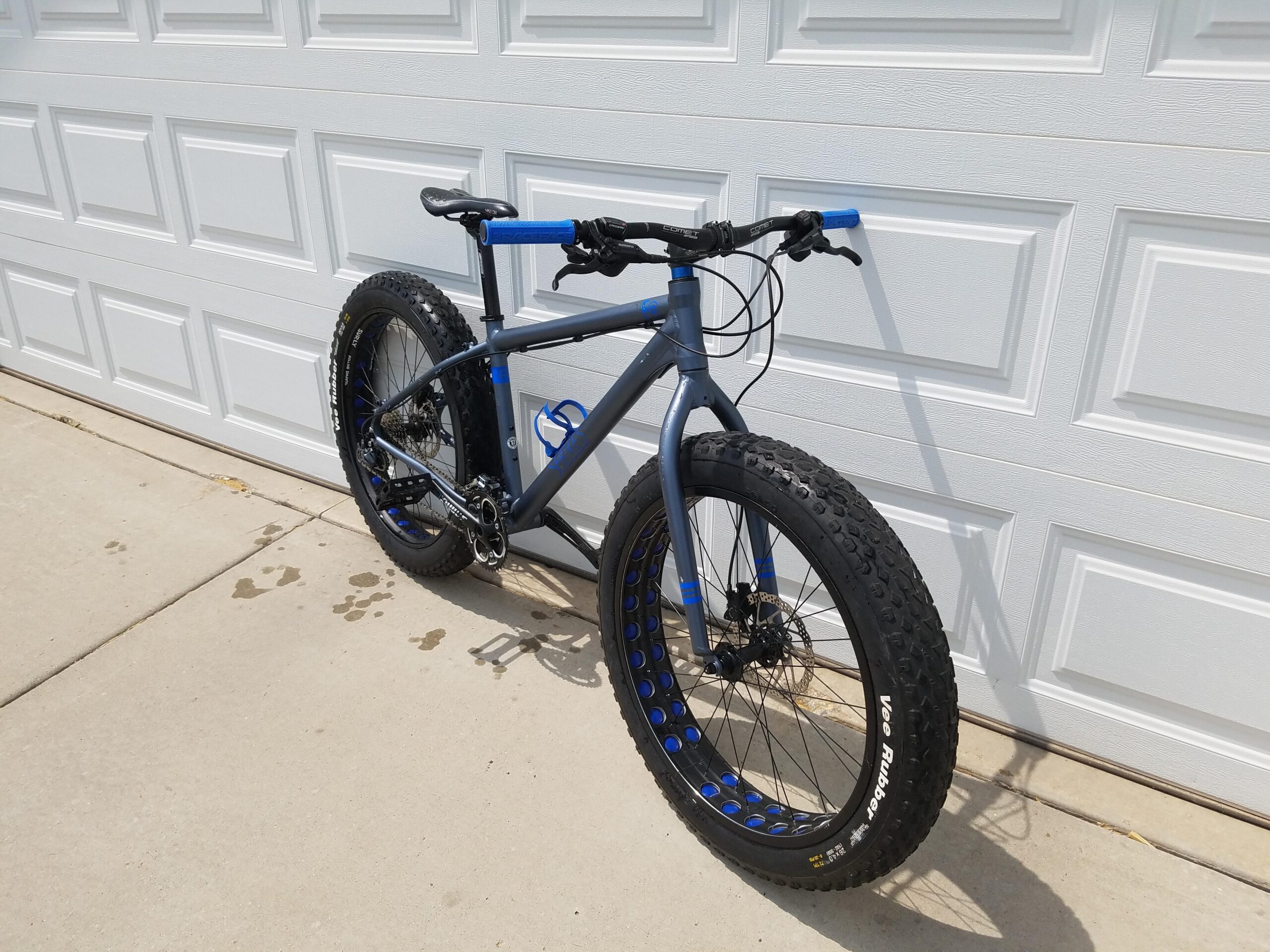 SE F@r: A gray and blue fat bike leaning against a white garage door, featuring large, knobby tires designed for off-road riding. The bike is positioned on a concrete surface with some water spots nearby.