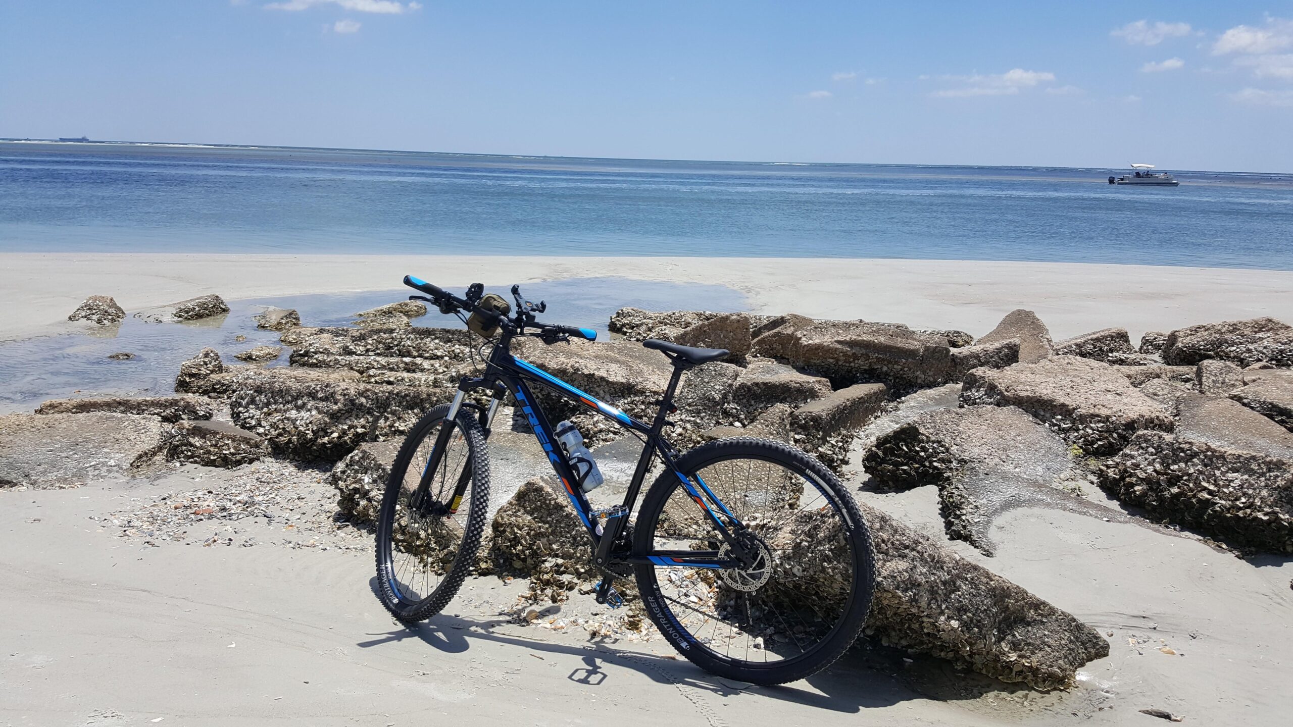 Trek Marlin 7: A mountain bike stands on a sandy beach beside rocky formations, with calm blue waters and a clear sky in the background.