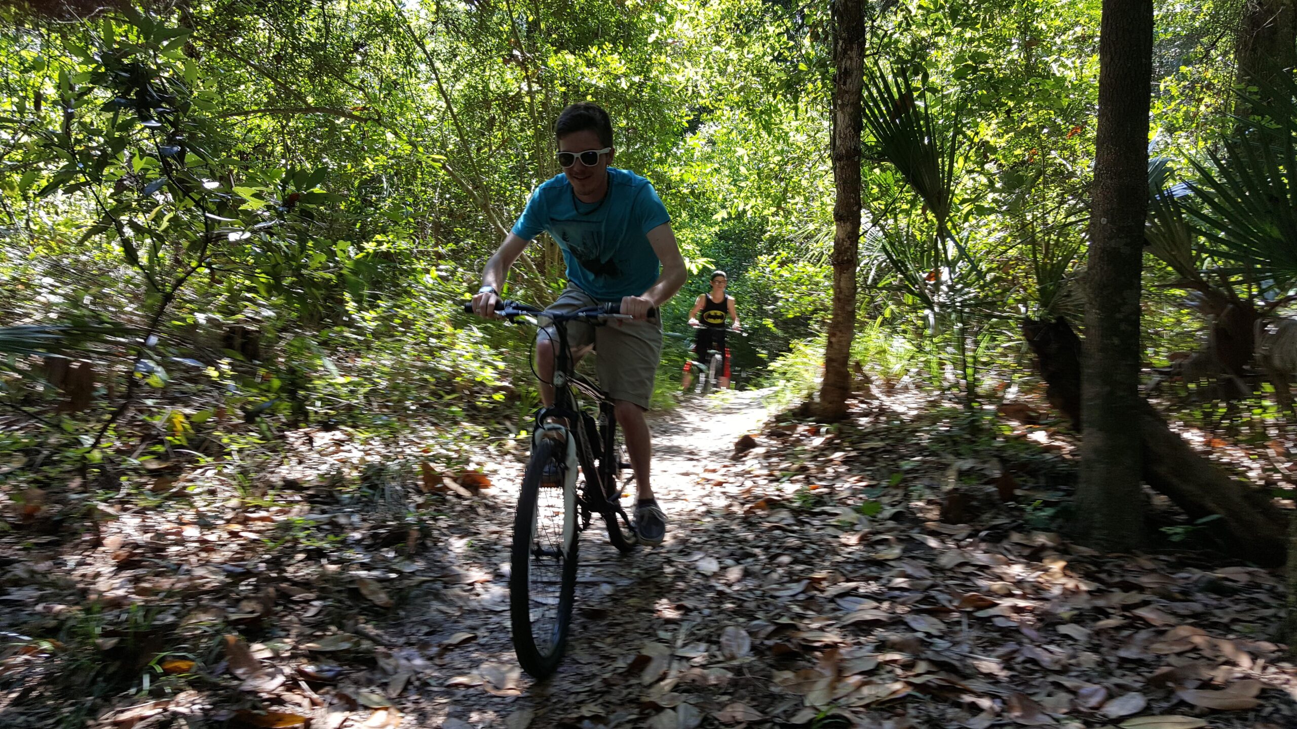 Two individuals are riding mountain bikes along a narrow trail in a lush, green forest. One rider, wearing a blue t-shirt and sunglasses, is in the foreground, focused on navigating the path, while a second rider, dressed in a black tank top, follows behind. Sunlight filters through the trees, illuminating the foliage and scattered leaves on the ground. Kathryn Abby Hanna Park mountain bike trail.
