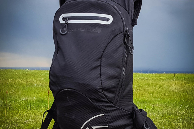 A black backpack with a mesh pocket displayed on a wooden post in a grassy field, under a cloudy sky. The backpack features a zipper pocket and a logo on the side.