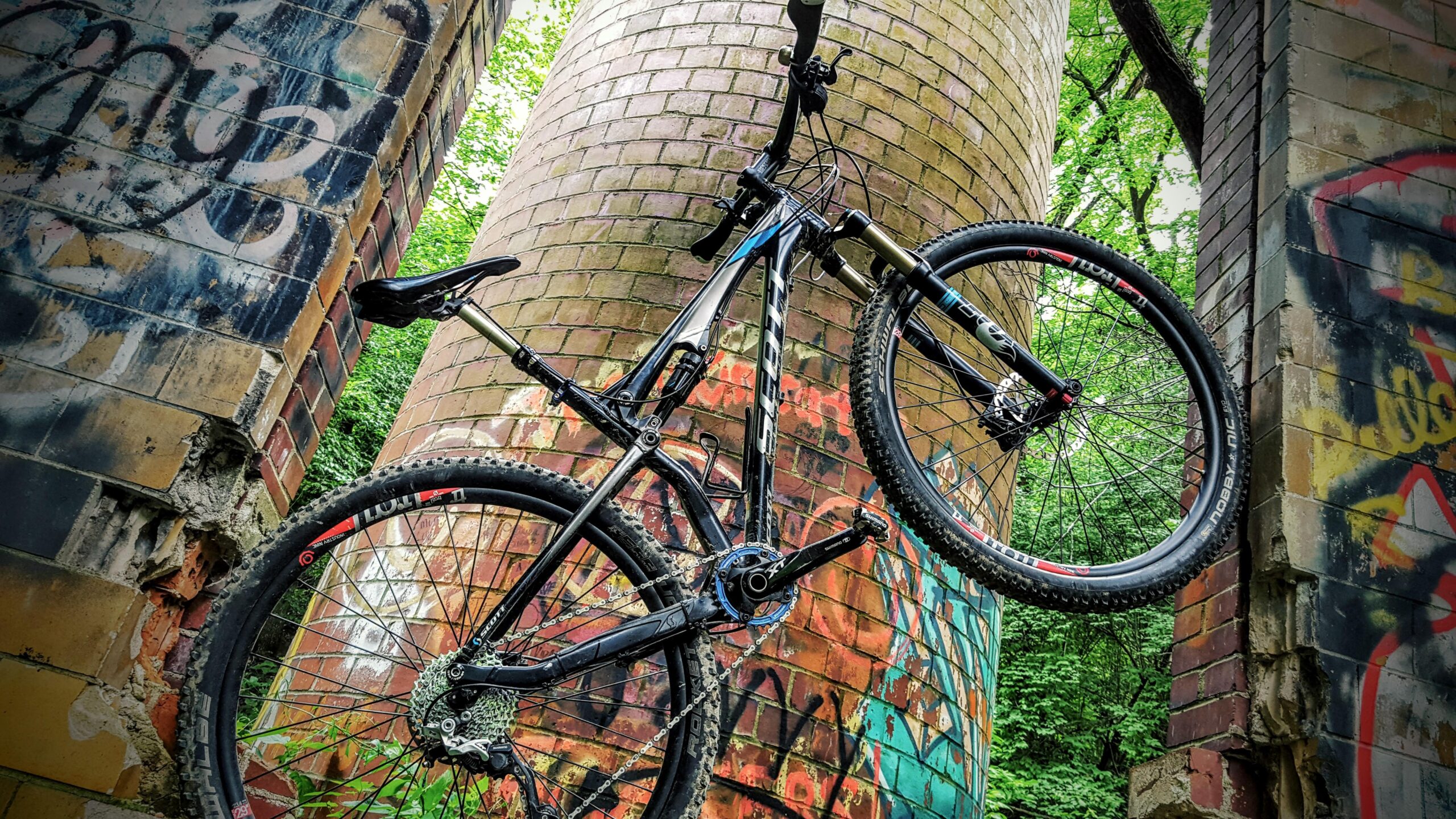 A mountain bike leaning against two weathered brick pillars covered in graffiti, surrounded by lush greenery. The bike is positioned with its front wheel elevated, showcasing its design and components. Devou Park mountain bike trail.