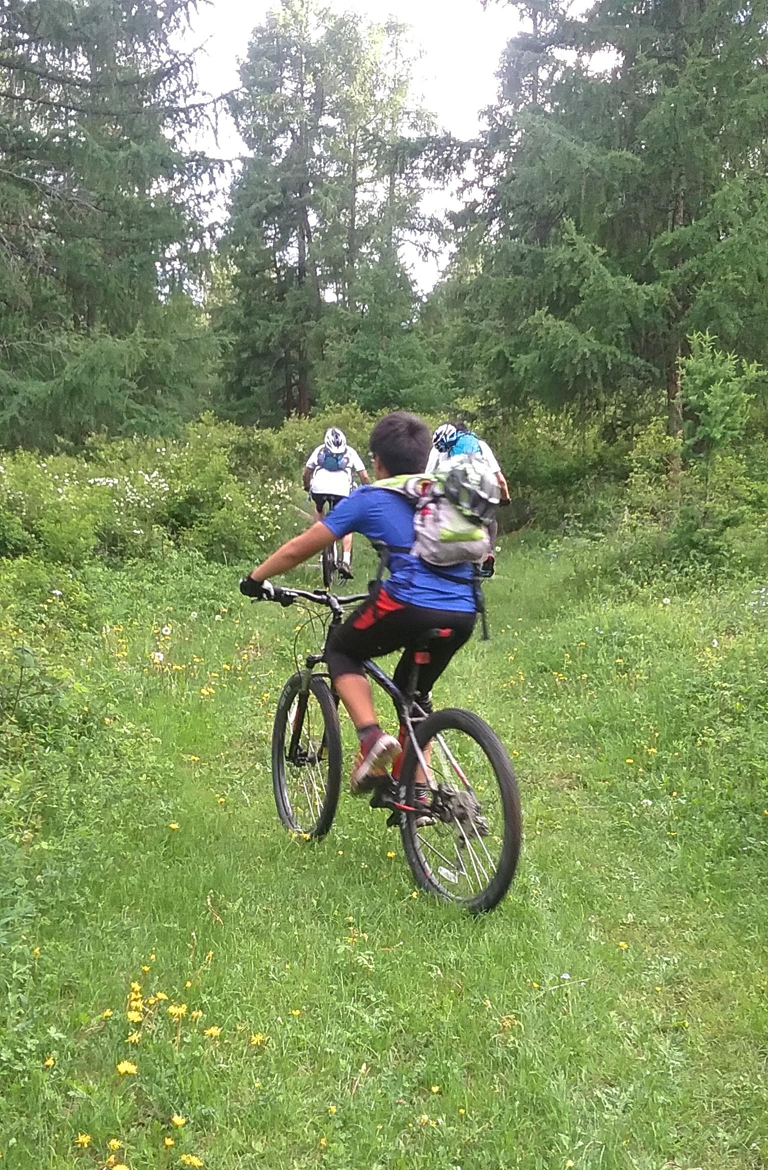 A child riding a mountain bike on a grassy trail in a forested area, with tall trees and wildflowers in the surroundings. Another cyclist is visible ahead, also navigating the trail. Chuluut Am mountain bike trail.