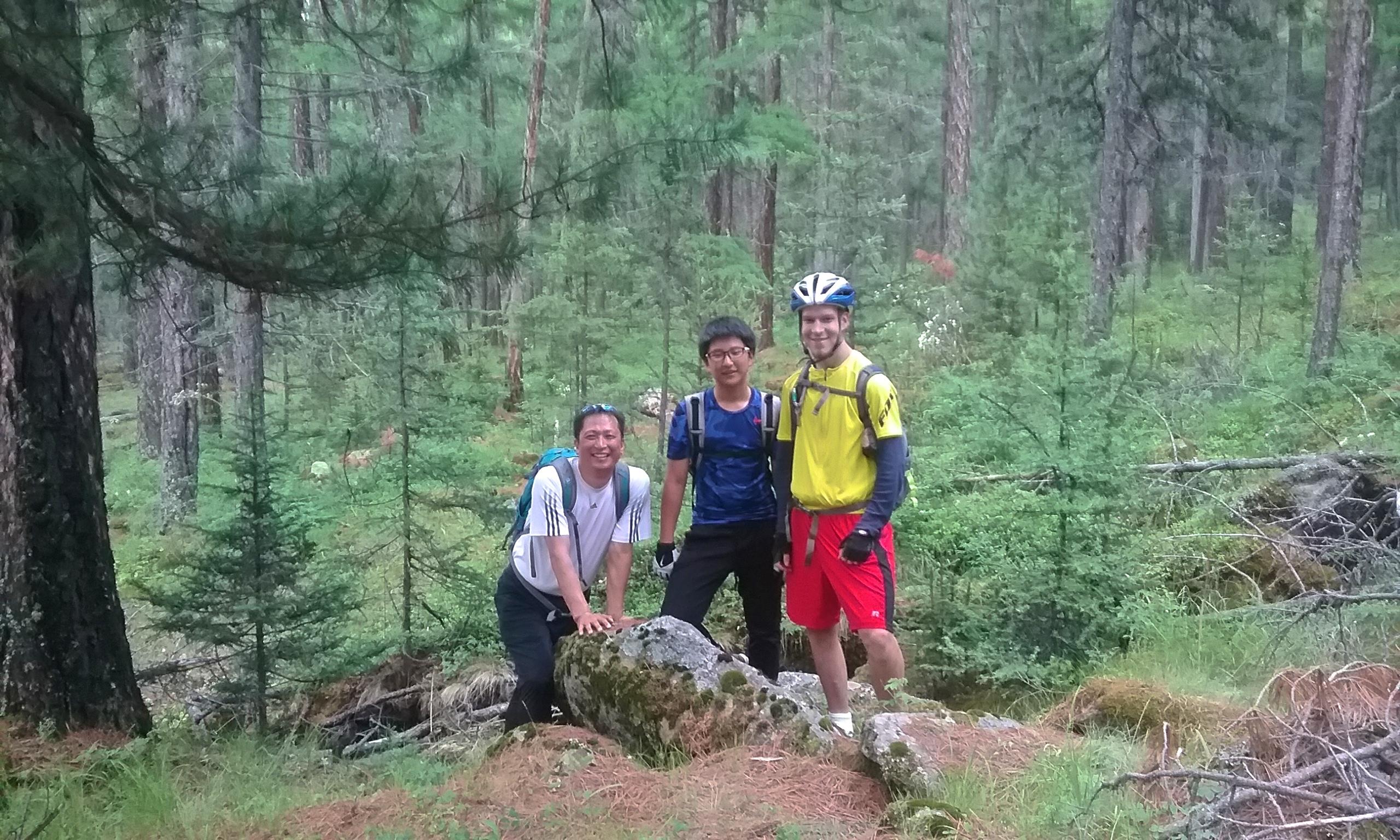 Three friends posing together on a moss-covered rock in a lush forest. The background features tall trees and greenery, indicating an outdoor hiking or biking adventure. Two individuals are wearing casual outdoor clothing, while one is dressed in biking gear, including a helmet. Chuluut Am mountain bike trail.