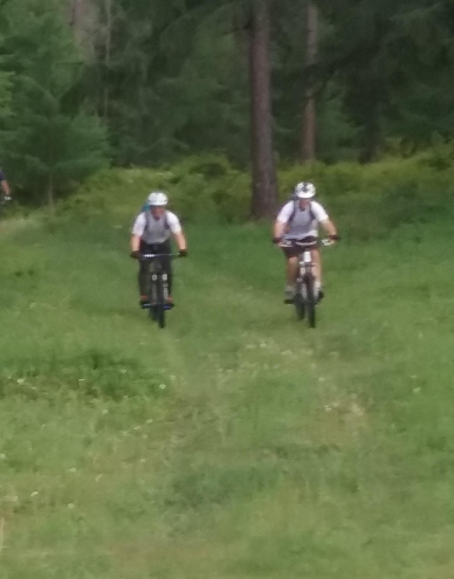 Two mountain bikers riding on a grassy path through a forested area, surrounded by trees and greenery. Chuluut Am mountain bike trail.