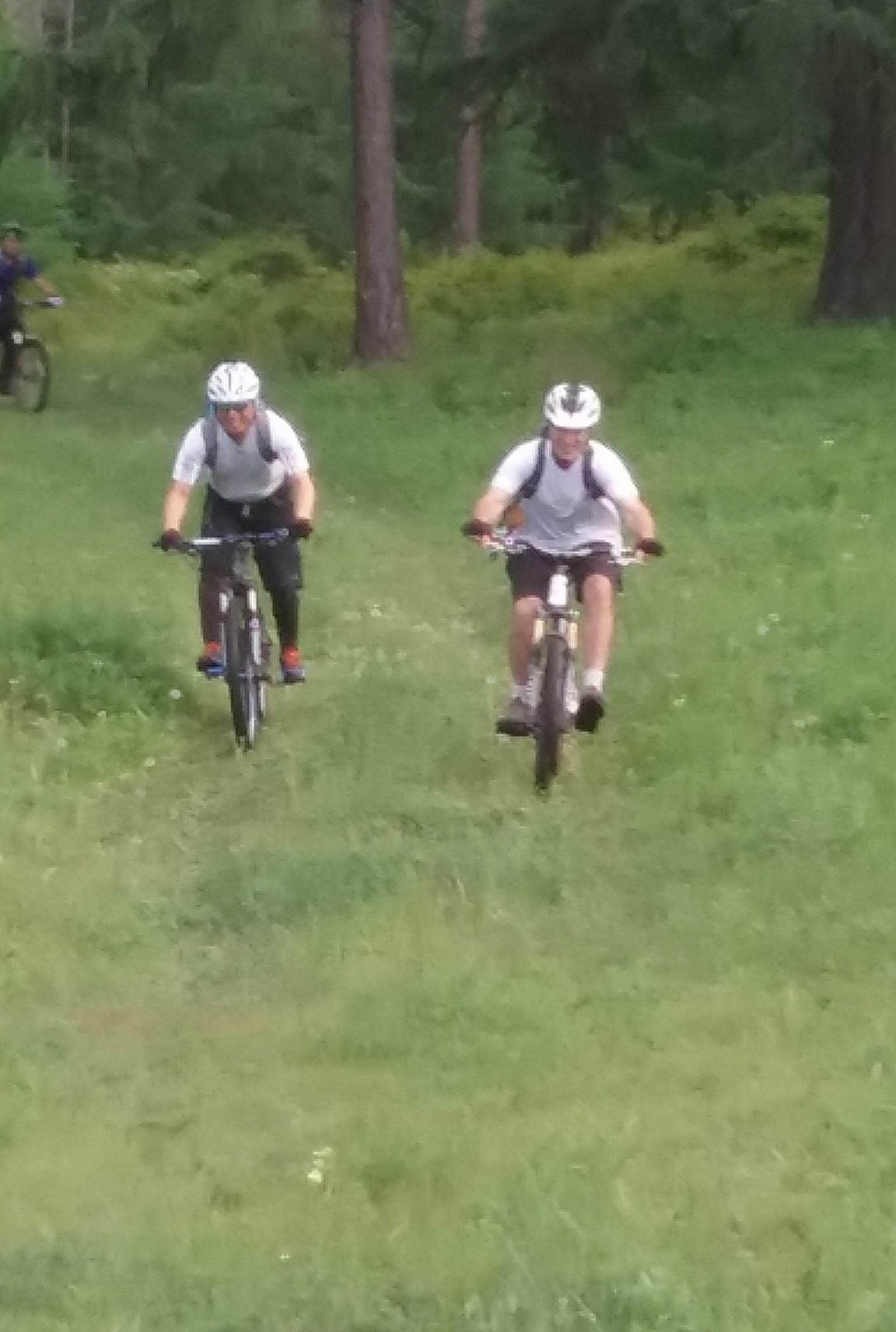 Two mountain bikers navigate a grassy trail surrounded by trees. Both riders are wearing helmets and athletic clothing, with one biker slightly ahead of the other. In the background, a third biker can be seen riding along the trail. The scene conveys a sense of outdoor adventure and enjoyment of nature. Chuluut Am mountain bike trail.