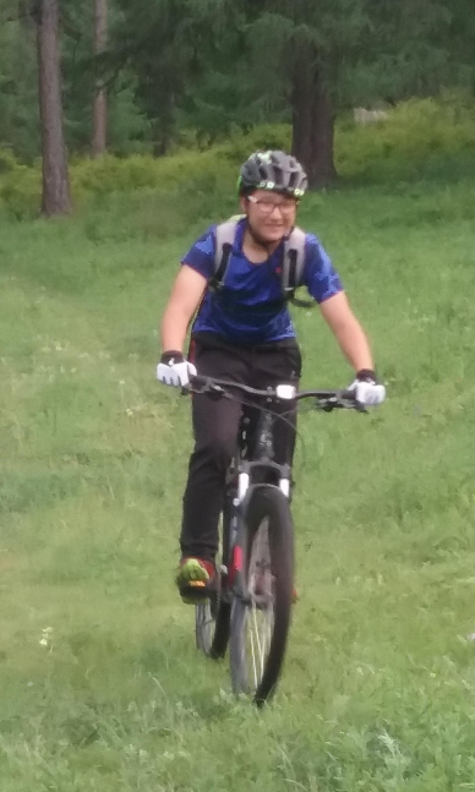 A person riding a mountain bike on a grassy trail in a wooded area, wearing a helmet and cycling gear. They appear to be smiling and enjoying the ride, surrounded by tall trees and greenery. Chuluut Am mountain bike trail.