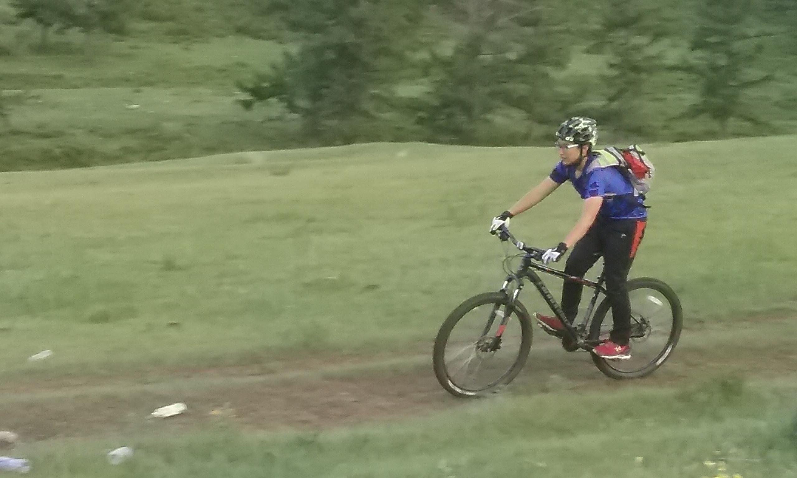 A person riding a mountain bike along a dirt path in a grassy landscape, wearing a helmet, gloves, and a blue shirt. Green hills and trees are visible in the background. Chuluut Am mountain bike trail.
