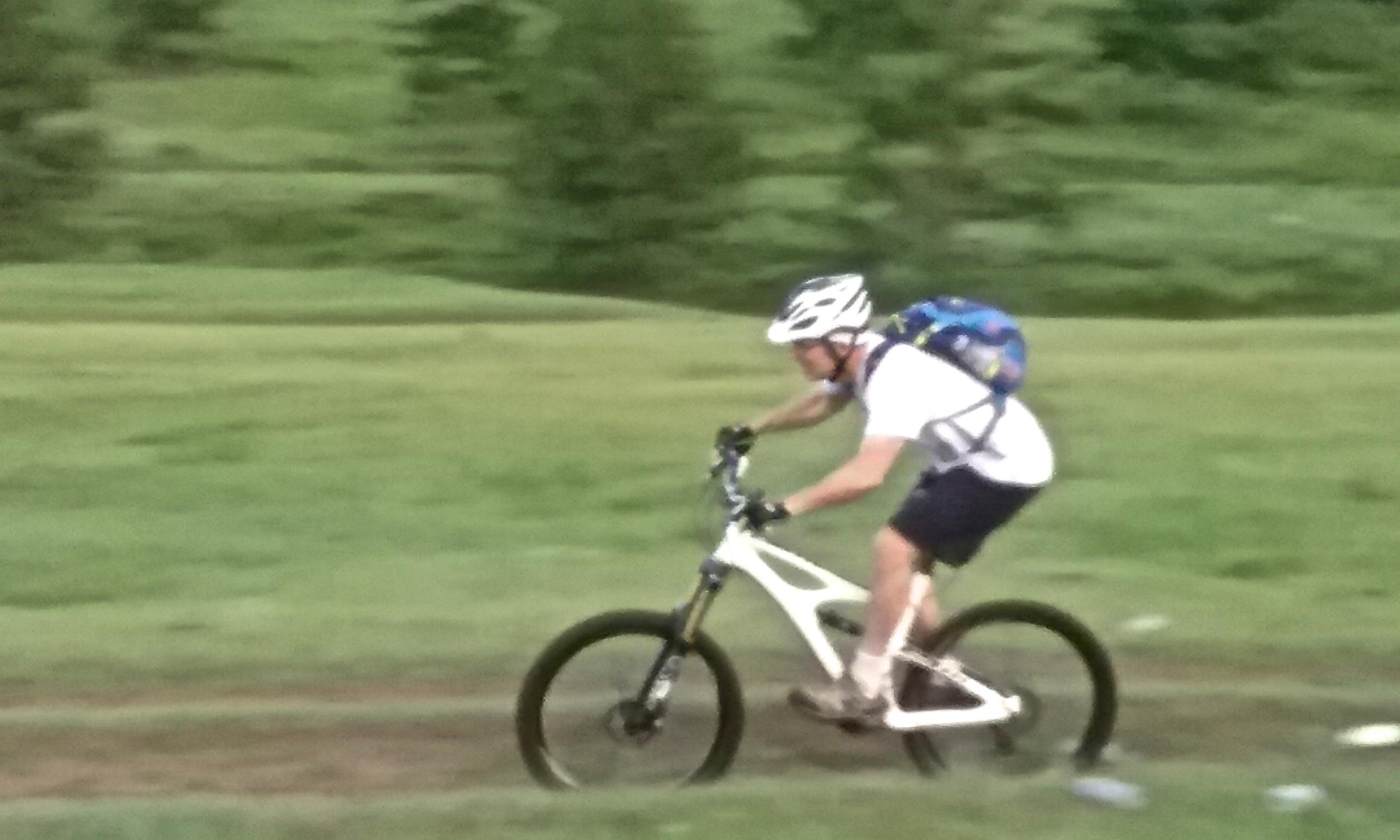 A person riding a mountain bike on a grassy terrain, wearing a white shirt, shorts, and a helmet, with a backpack. They are leaning forward as they navigate the path. Chuluut Am mountain bike trail.