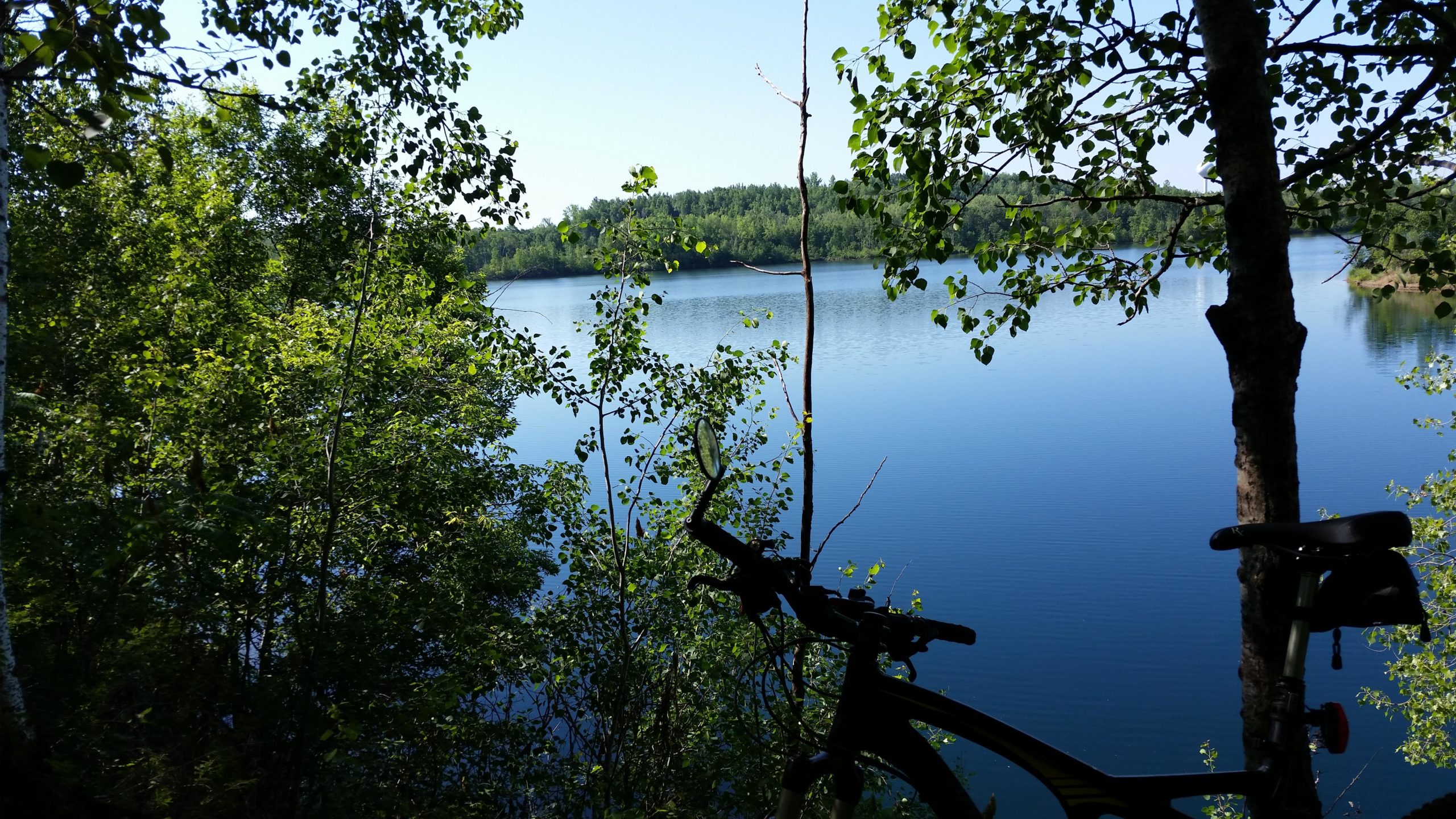 A serene view of a calm lake surrounded by lush greenery, with a bicycle parked in the foreground. The clear blue sky reflects on the water, creating a peaceful outdoor setting. Portsmouth unit/Dragline/Man High Hill mountain bike trail.