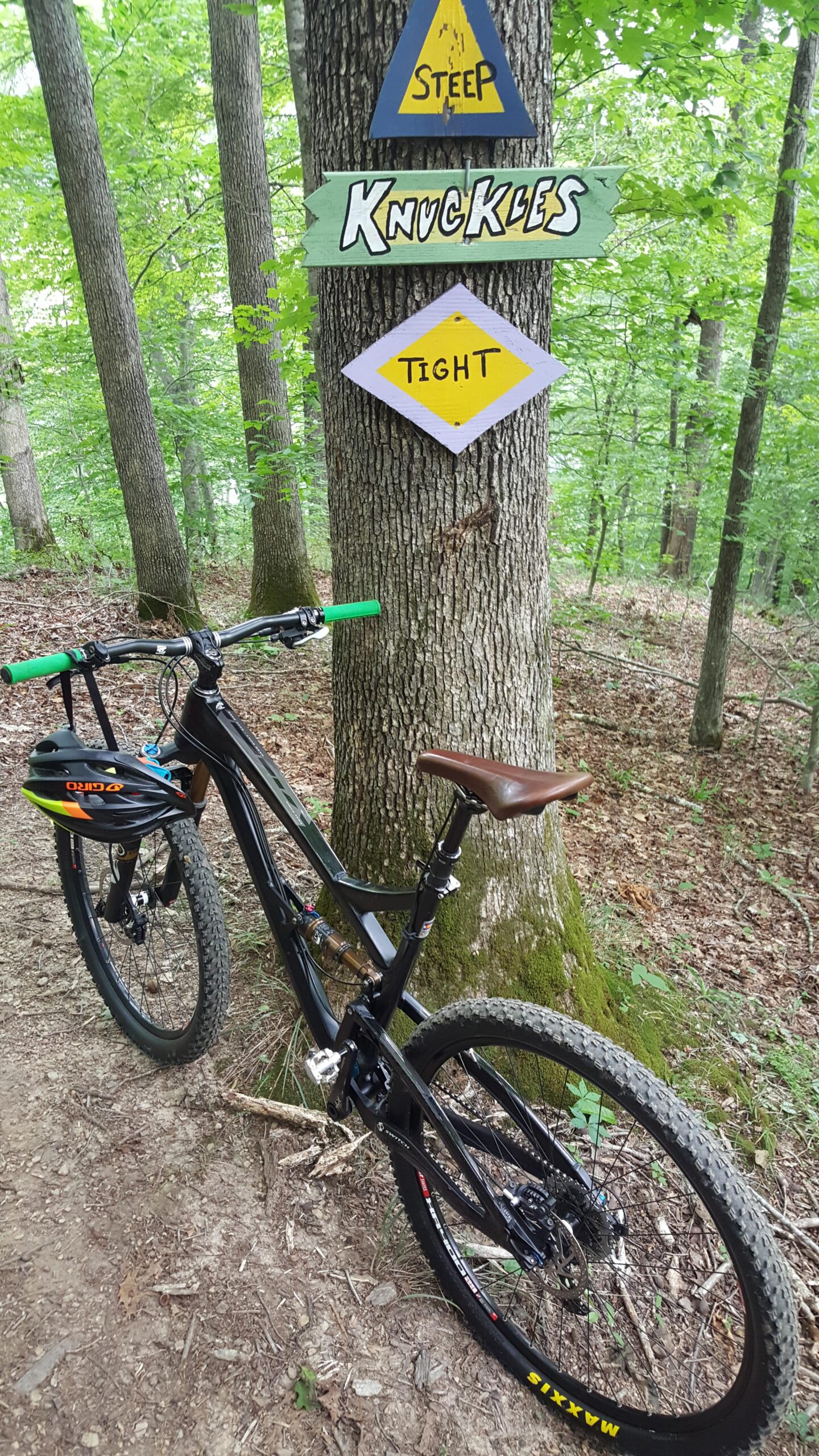 Yeti SB95: A mountain bike parked beside a tree, with trail signs indicating "Steep," "Knuckles," and "Tight" in a wooded area. The bike features green handlebars and a brown saddle, surrounded by lush green foliage and a dirt path.