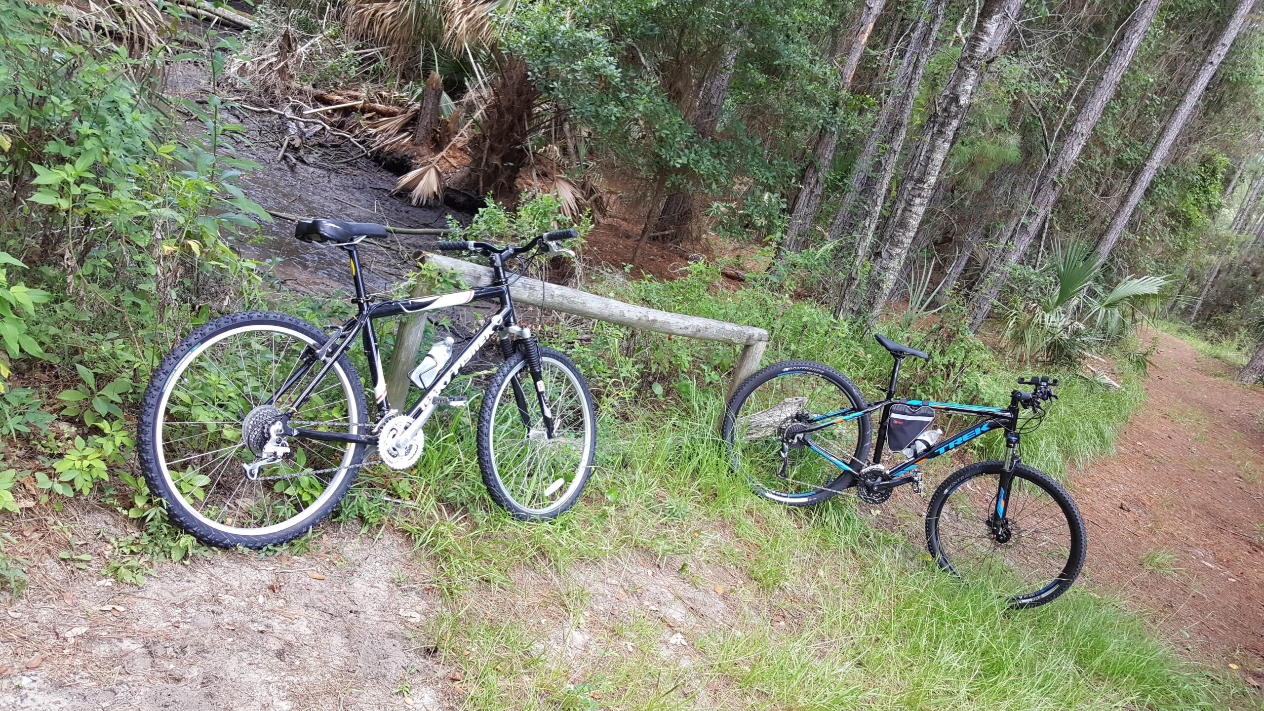 Gary Fisher Wahoo: Two mountain bikes leaning against a wooden fence in a lush forest setting, with a small stream visible in the background. The scene features dense greenery, tall trees, and dirt trails, suggesting an outdoor recreational area for biking.