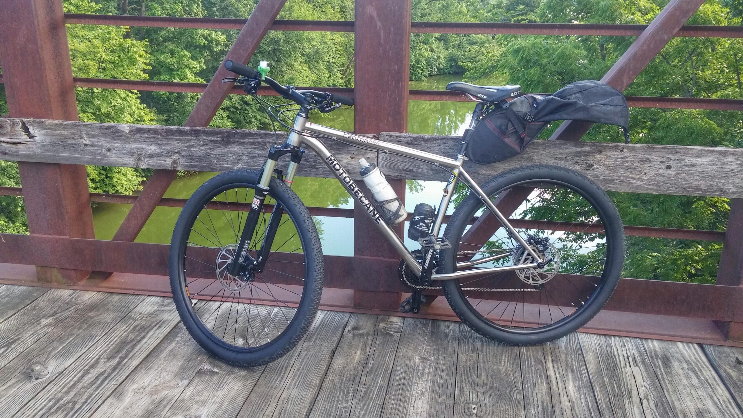 Motobecane Fantom PRO Titanium: A mountain bike with a water bottle attached rests on a wooden bridge railing, surrounded by lush green trees and a reflective body of water below. A small black bag is secured on the bike's rear. The bridge features a combination of wooden and metal elements.