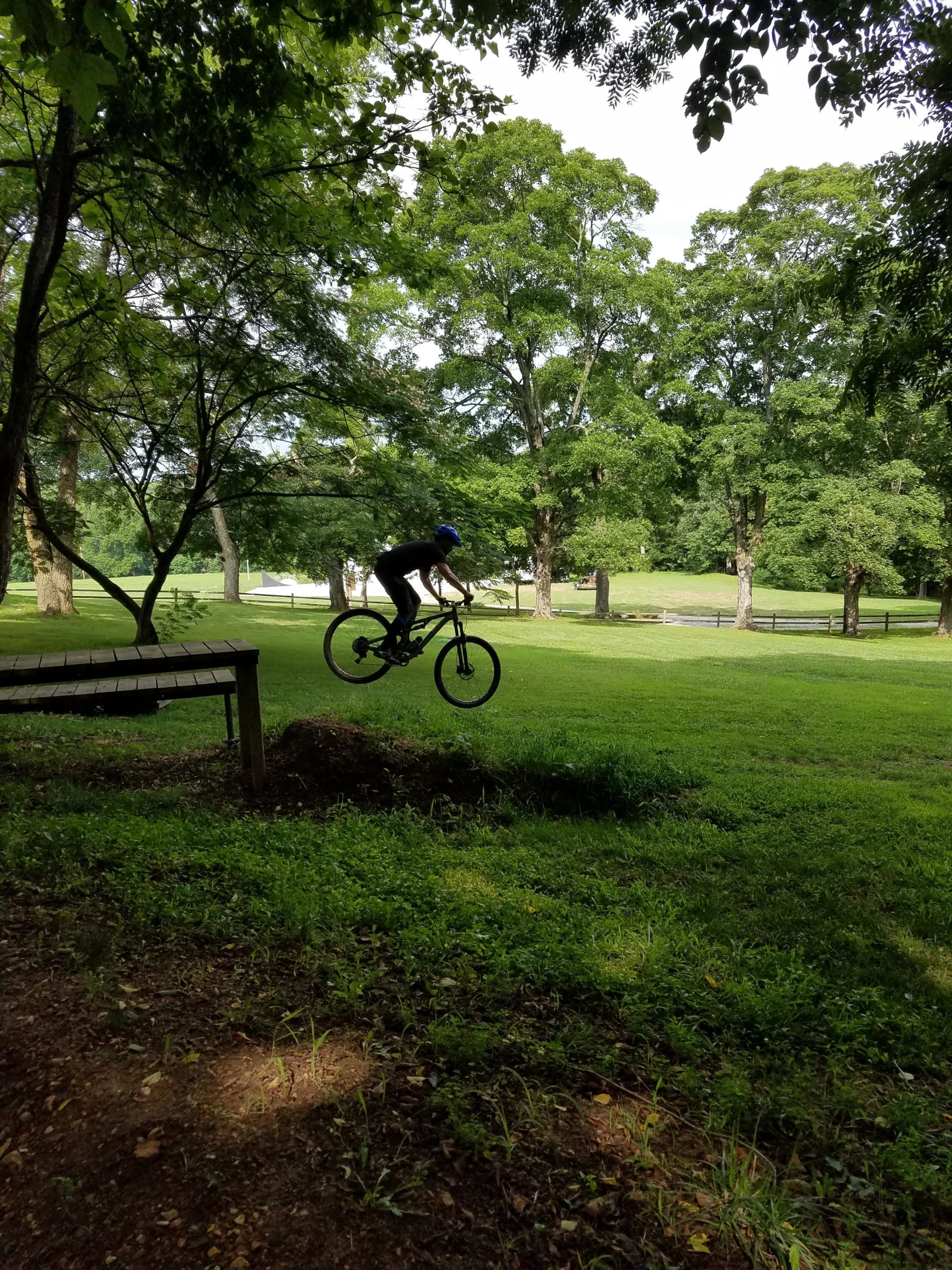 A person riding a mountain bike is jumping over a small hill on a grassy landscape, surrounded by tall trees and a clear blue sky. The scene captures a lively outdoor activity in a natural setting. Two Rivers Bike Park mountain bike trail.