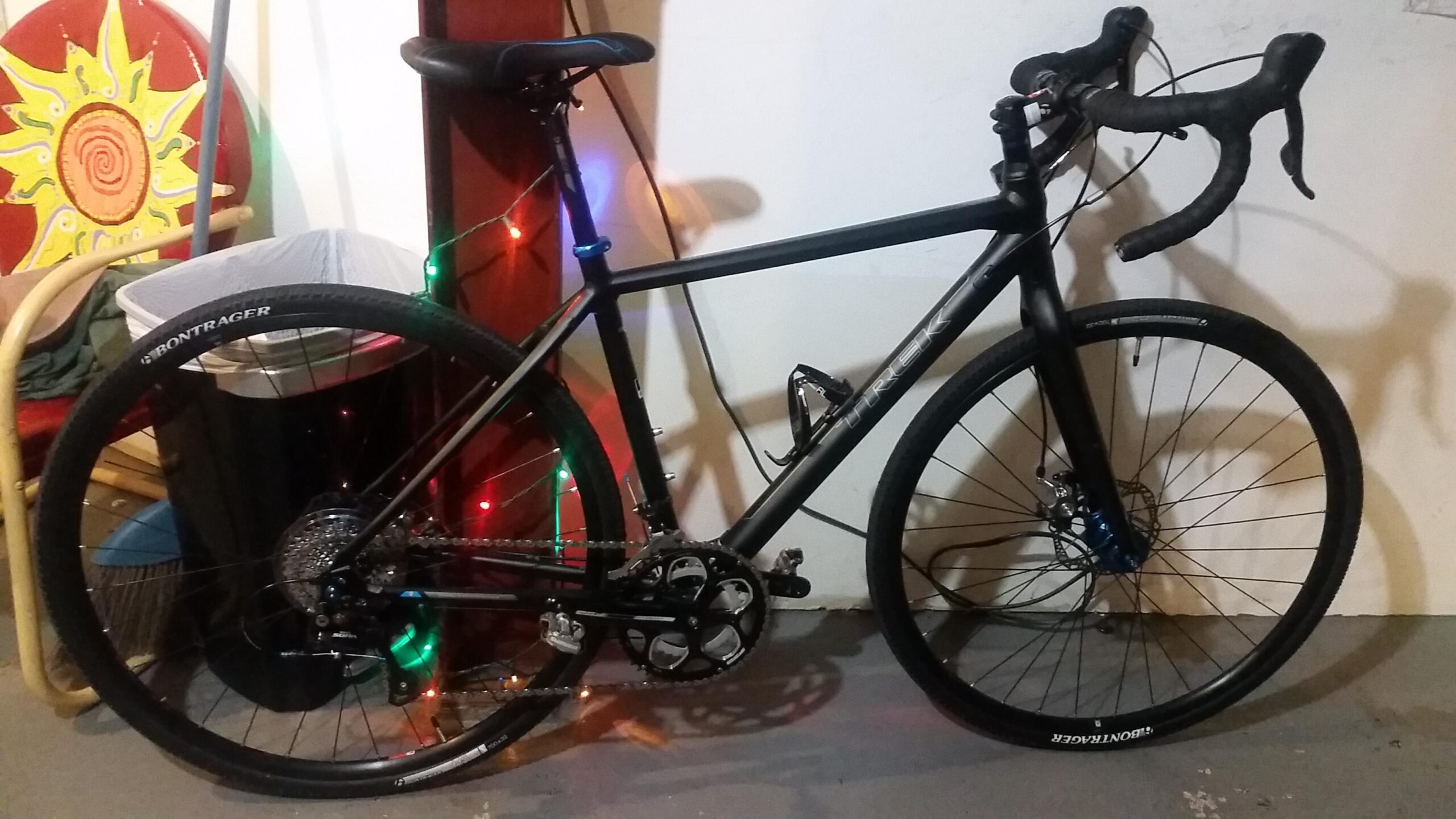 Trek Crossrip Elite: A black Trek bicycle parked in a garage, featuring drop handlebars, disc brakes, and Bontrager tires. In the background, there is a colorful sun-painted chair and a trash bin, along with some decorative lights on the wall.