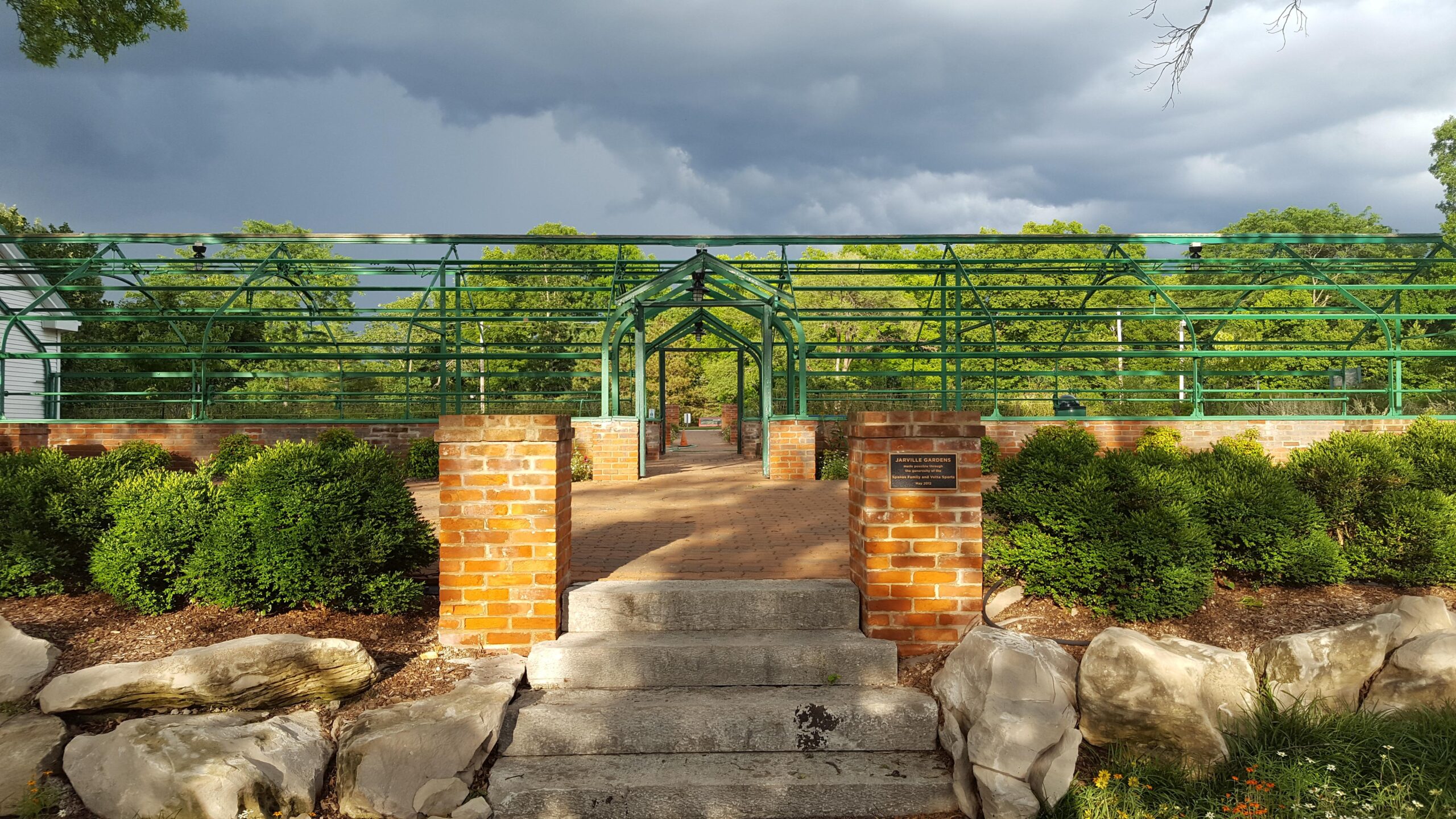 A landscaped path leading through a green metal archway, flanked by neatly trimmed shrubs and stone edging. The scene is set against a backdrop of dark, cloudy skies, creating a contrast with the surrounding greenery. The path is made of brick and leads into a garden area, with a sign indicating the location as "Asheville Gardens." Queeny Trails mountain bike trail.