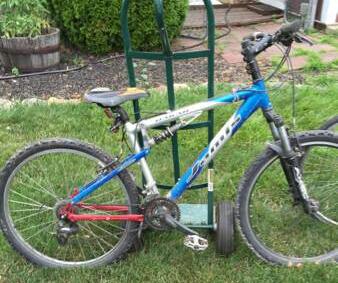 Jamis Dakar XC: A blue and silver mountain bike with a red rear derailleur, resting on a patch of grass beside a green cart. The bike appears to be used, with some dirt on the tires and frame.