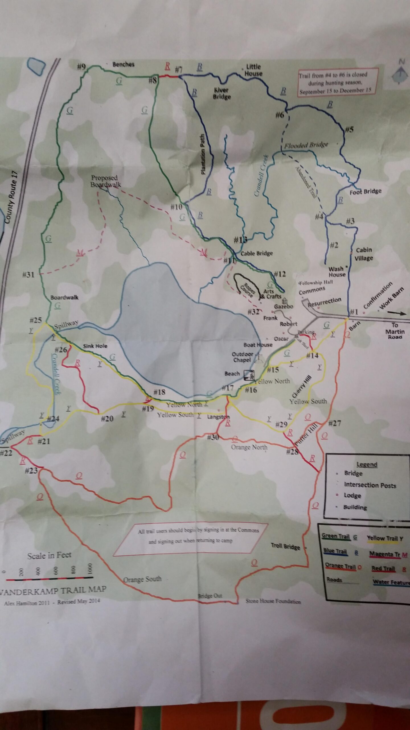 A color-coded trail map of Vanderkamp, indicating various hiking paths, bridges, and notable locations such as cabins and a beach. The map features a legend for trail types (green, yellow, blue, orange, and red) and includes labels for trail markers and features like bridges and lodges. Important notes about trail access and seasonal closures are also highlighted. Vanderkamp mountain bike trail.