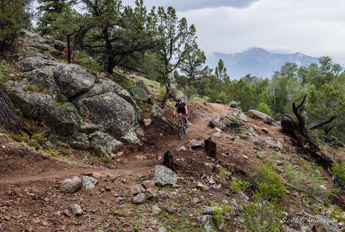 A mountain biker navigating a dirt trail surrounded by rocky terrain and trees, with distant mountains and cloudy skies in the background. Midland Hills Trails mountain bike trail.