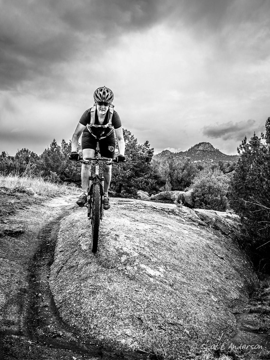 A mountain biker navigating a rocky trail with a determined expression, set against a backdrop of cloud-covered mountains and trees. The image is in black and white, emphasizing the rugged terrain and dynamic movement. Midland Hills Trails mountain bike trail.