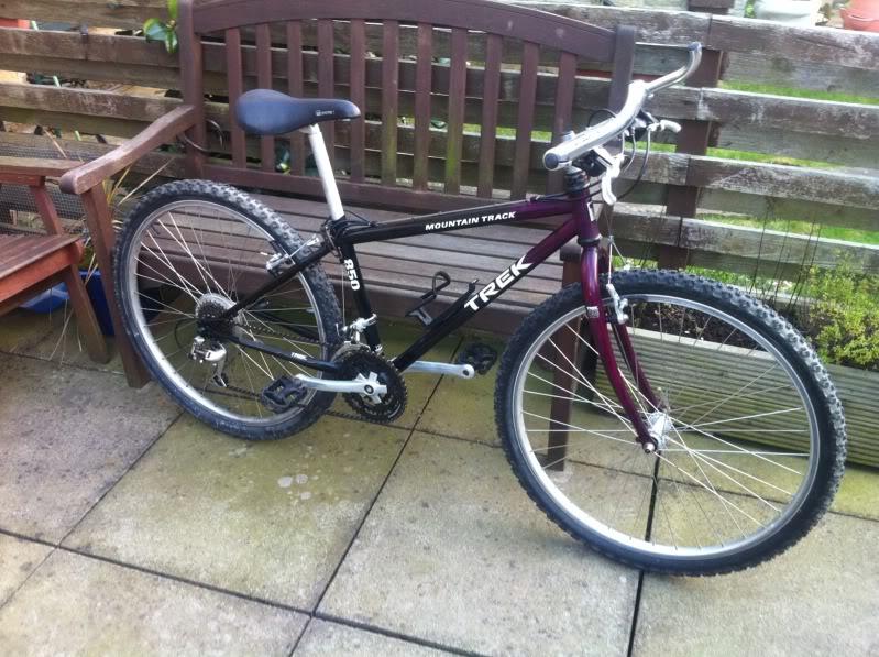 Trek Trek 850 Mountain Track: A black and maroon Trek Mountain Track bicycle is leaning against a wooden bench on a patio. The bike has thick, knobby tires suitable for off-road use and a simple gear system. The background features a wooden fence and potted plants, creating a cozy outdoor setting.