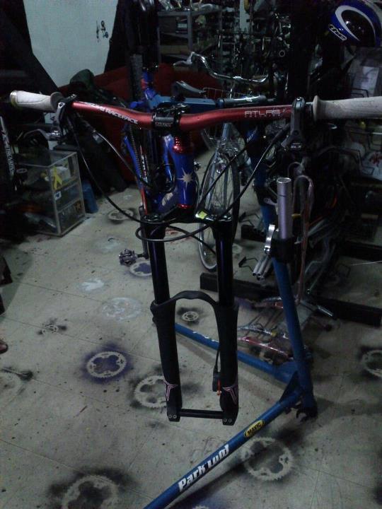 On-One 456: A bicycle handlebar and front fork mounted on a repair stand, surrounded by various bike tools and parts in a workshop setting. The handlebars feature white grips and a red decorative finish, while the bike frame is visible in the background along with other bicycles. The floor has markings and stains indicative of a working environment.