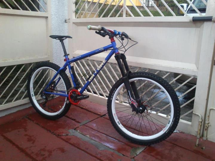 On-One 456: A blue mountain bike with a black front fork, red accents, and white wheels, positioned on a tiled surface near a partially open gate. The bike features a sleek design suitable for off-road riding.
