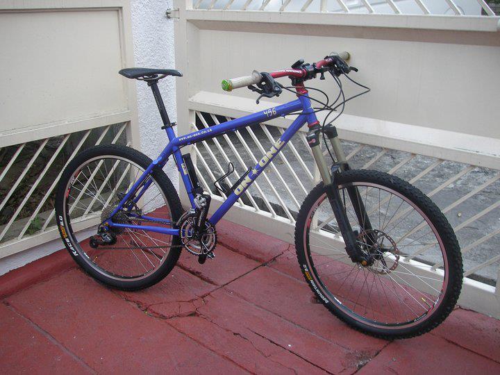 On-One 456: A blue mountain bike with a metallic frame, featuring thick tires and suspension forks, parked on a red tiled surface. The bike is positioned near a white fence.