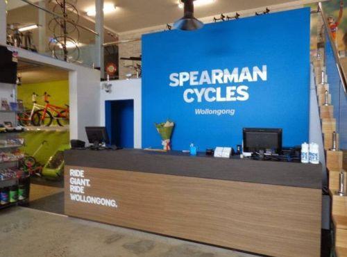 Interior of Spearman Cycles in Wollongong, featuring a reception area with a blue wall displaying the store name. The counter is made of wood and has a flower arrangement and electronic devices on it. Bicycles are visible in the background, and there