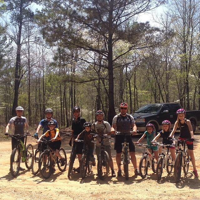 A group of eight adults and children stands together on a dirt path, all wearing helmets and posing with their bicycles. They are surrounded by trees in a forested area on a sunny day. A pickup truck is parked in the background. The group includes a mix of genders and ages, showcasing a shared enthusiasm for mountain biking. Sylaward mountain bike trail.