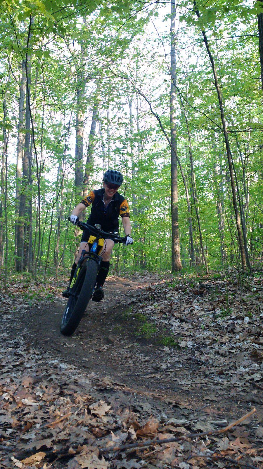 A person riding a mountain bike on a forest trail, surrounded by tall green trees. The rider is leaning forward, navigating a bend on the trail, with sunlight filtering through the leaves. The ground is covered in fallen leaves and twigs, showcasing a vibrant natural setting. Dufferin County mountain bike trail.