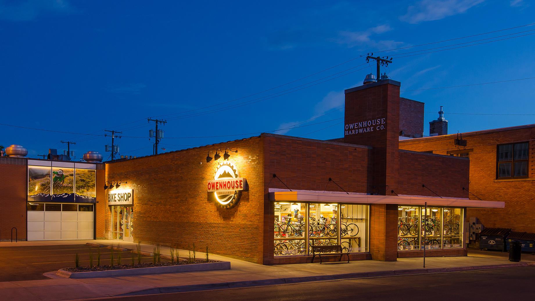 A brick building with illuminated signage reading "Owenhouse" at dusk, featuring a bike shop on the left with large windows showcasing bicycles inside. The right side of the building displays "Owenhouse Hardware Co." in large lettering, with parking spaces in front and utility poles in the background. The scene conveys a welcoming atmosphere with well-lit storefronts.